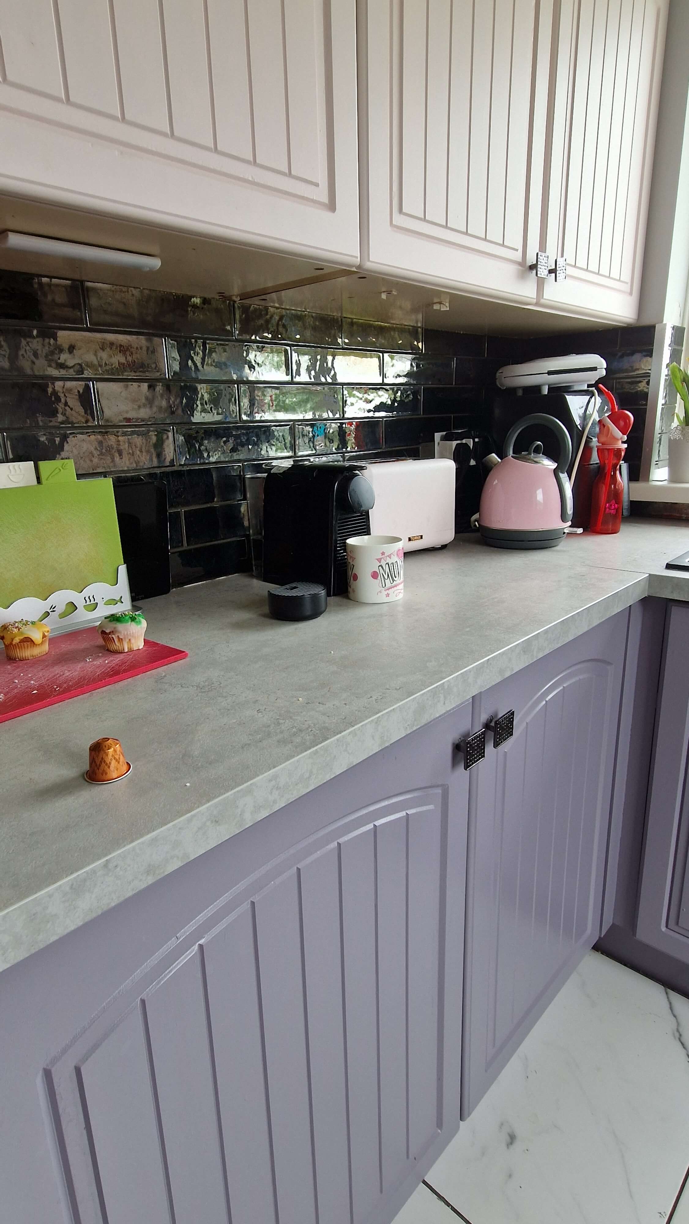  kitchen with concrete effect countertop, black, chrome effect wall tile, light pink and lavender kitchen cupboards. There is marble carrera tiles on the floor